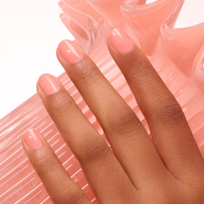 A hand with medium brown skin displays neatly manicured nails painted in Manucurist Glacé, an ultra-glossy light pink, resting on a translucent, textured pink object with curved ridges.