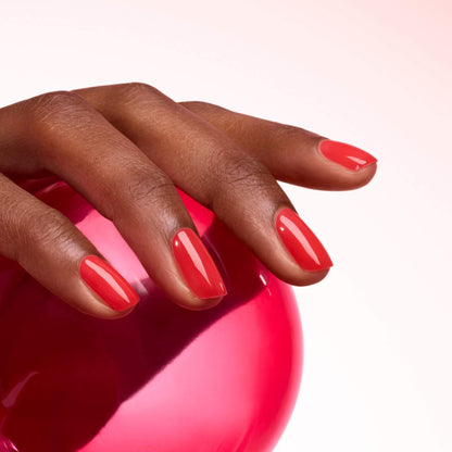 A hand with neatly manicured, ultra-glossy Twizzle red nails by Manucurist rests on a shiny, pinkish-red sphere against a soft gradient background.