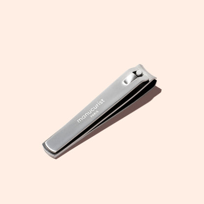 A silver Manucurist Nail Clipper, designed for precise manicures and ideal for shaping natural nails, is engraved with the brand name and displayed on a light beige background.