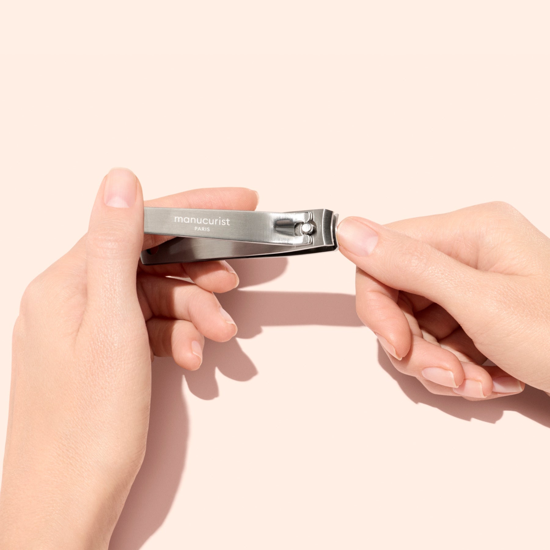 A person holds the Manucurist Nail Clipper in one hand and gets ready to trim the natural nails of the other against a pale background, highlighting a simple at-home manicure routine.