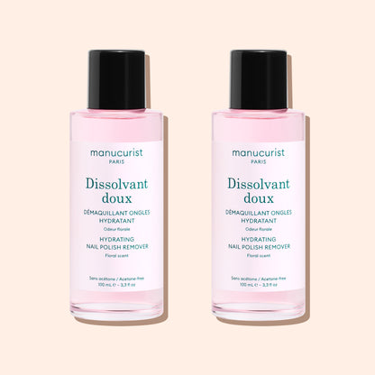 Two bottles of Manucurist Gentle Remover, an acetone-free nail polish remover with pink liquid, are side by side on a beige background. Each has a black cap and white label with text.