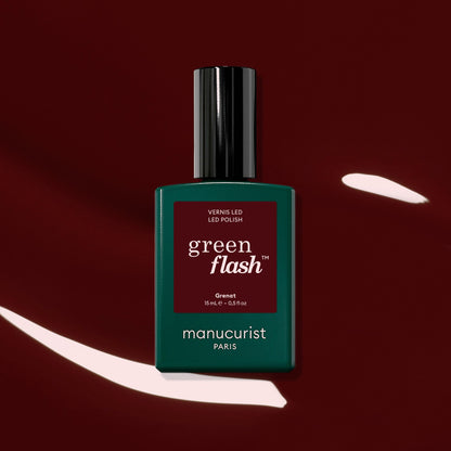 A bottle of Manucurist Grenat gel polish, set against a glossy earthy red nail polish background with white highlights.