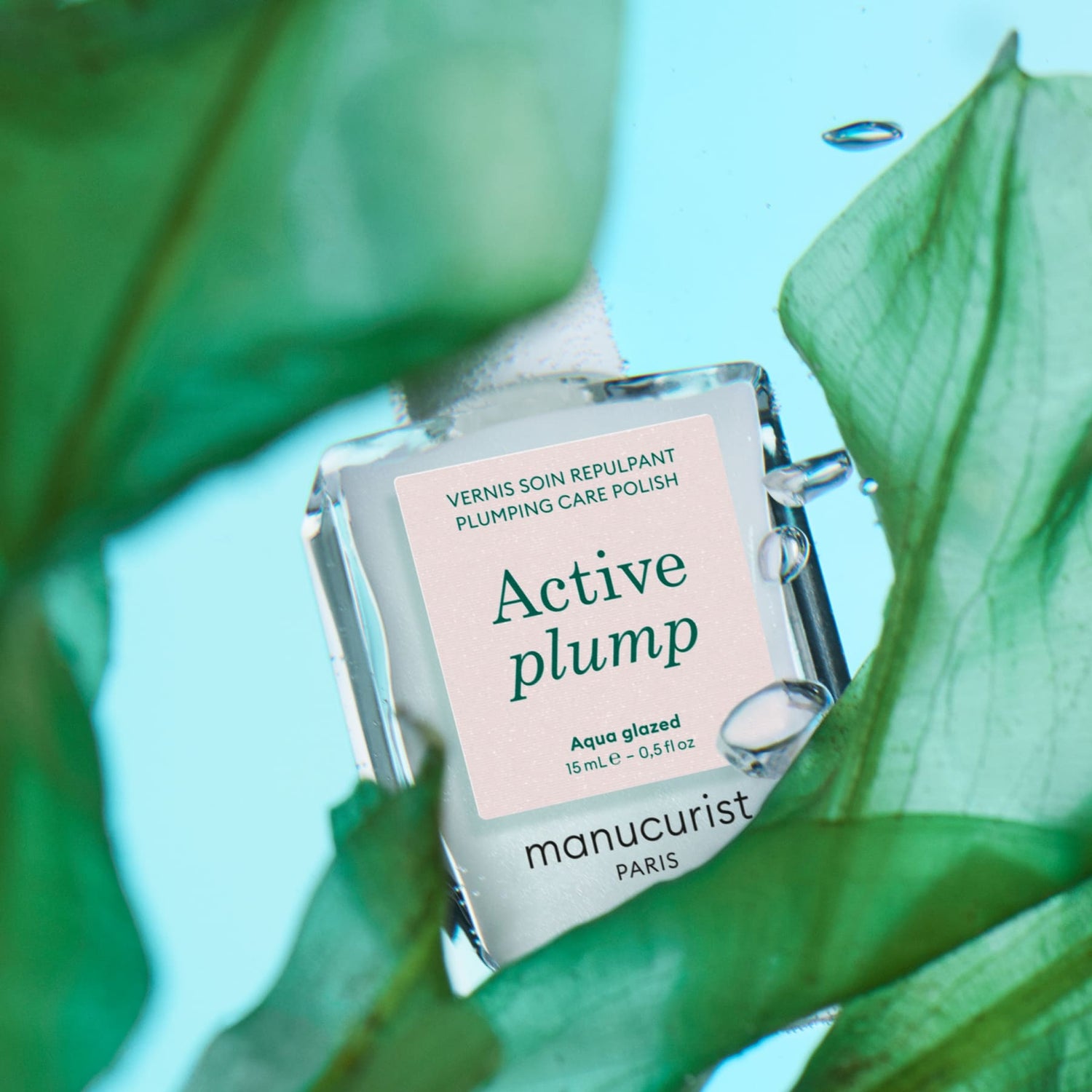 Manucurist Active Plump Aqua Glazed nail polish is shown among green leaves with water droplets on both, set against a light blue background, highlighting the formula’s hydrating benefits for nails.