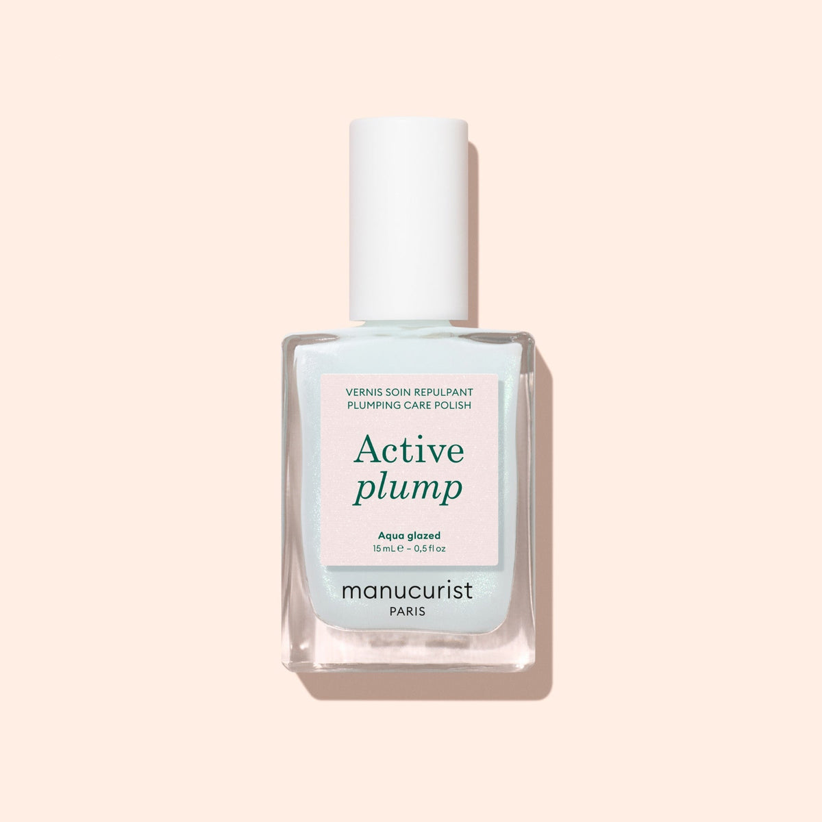 A bottle of Manucurist Active™ Plump Aqua Glazed nail polish with a white cap rests on a light pink background. The pale label features teal and pink text, reflecting the promise of hydrated nails and a luminous, glazed finish.