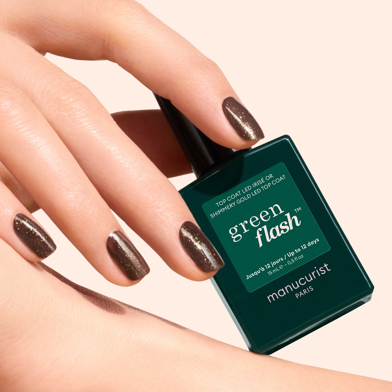 A hand with brown, shimmery nails featuring gold and silver shimmer holds a bottle of Manucurist Glitter Top Coat gel polish against a light background. The dark green bottle displays product information in white text.