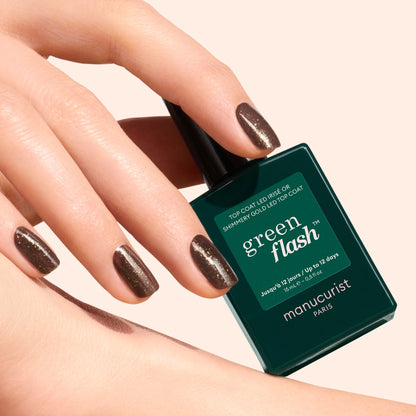 A hand with brown, shimmery nails featuring gold and silver shimmer holds a bottle of Manucurist Glitter Top Coat gel polish against a light background. The dark green bottle displays product information in white text.
