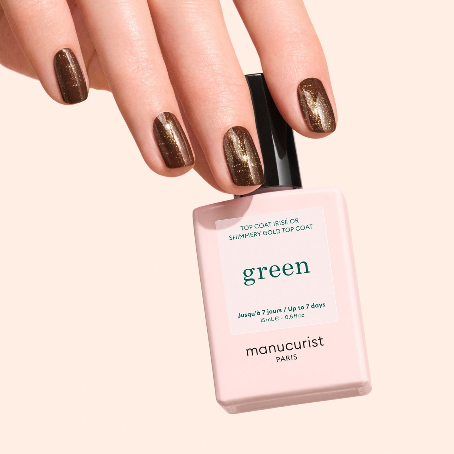 A hand with brown, shimmery nail polish holds a pale pink bottle of Manucurist Glitter Top Coat against a light beige background, showcasing its sparkling gel-effect finish.