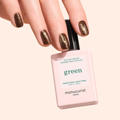 A hand with brown, shimmery nail polish holds a pale pink bottle of Manucurist Glitter Top Coat against a light beige background, showcasing its sparkling gel-effect finish.