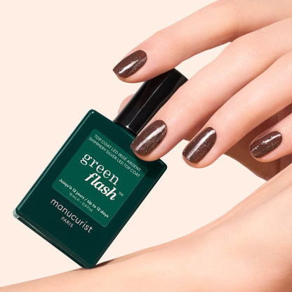 A hand with neatly manicured, dark brown glittery nails featuring gold and silver shimmer holds a green bottle of Manucurist Glitter Top Coat against a light beige background.