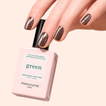 A hand with short, neatly manicured nails painted in shimmery brown holds a bottle of Manucurist Glitter Top Coat against a soft beige background. The gel-like finish adds subtle green glitter for extra shine.