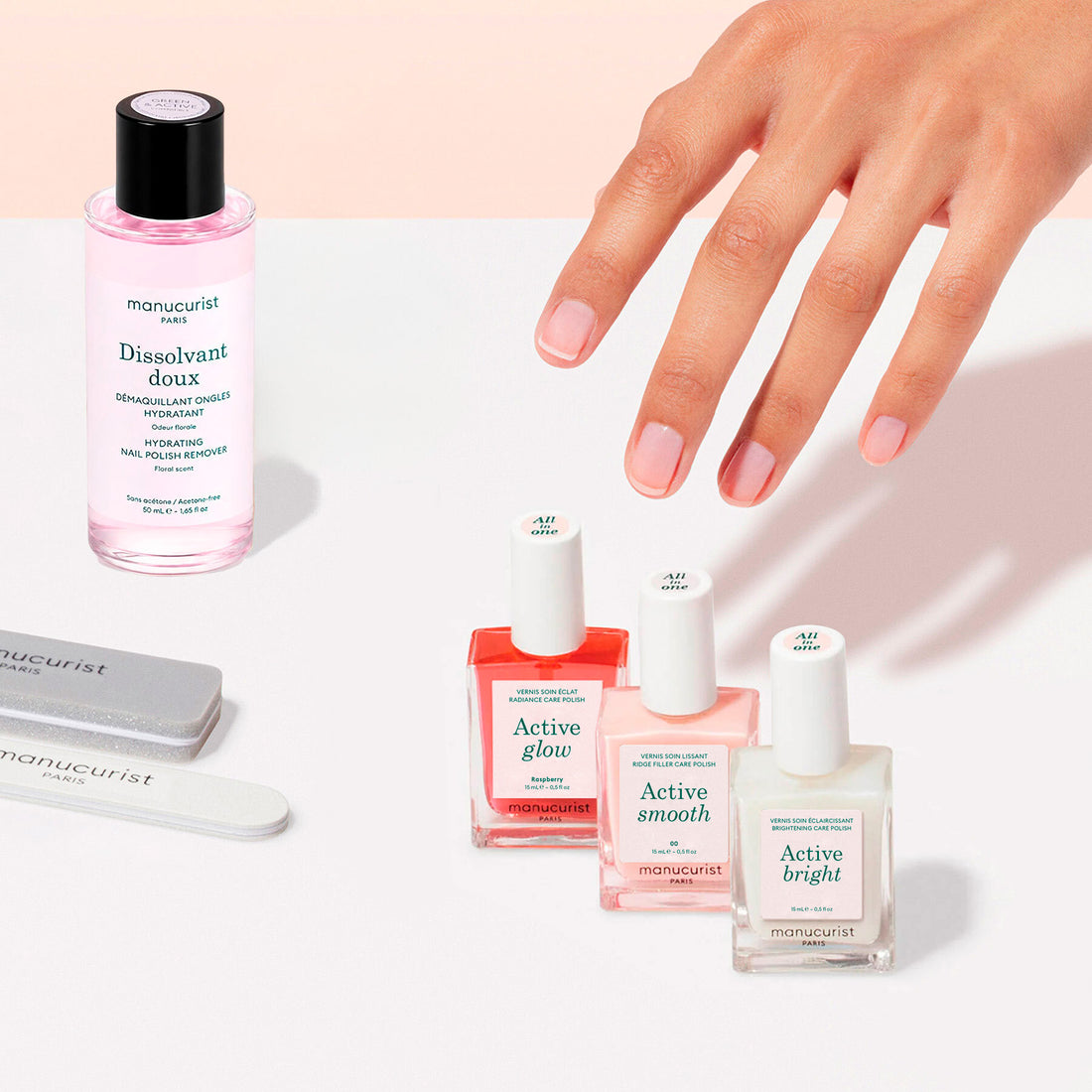 A hand poses near the Manucurist Active™ Manicure Kit, featuring three nail polishes (Active Glow, Active Smooth, and Active Bright), a pink nail polish remover, two nail files, and a nourishing nail serum on a white surface with a light pink background—perfect essentials for any nail care routine.