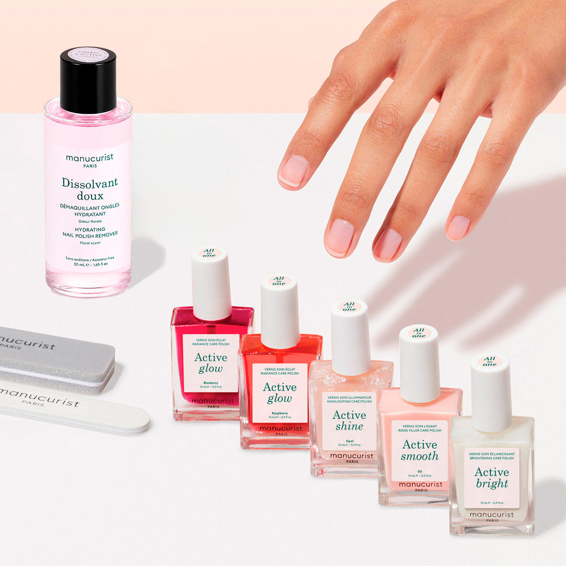 A hand with manicured nails reaches toward the Manucurist Active™ Super Manicure Kit, featuring five bottles of nail polish and a pink nail polish remover—all branded by Manucurist—with two nail files nearby on a pink and white background, creating the perfect setting for an indulgent or rejuvenating at-home manicure.