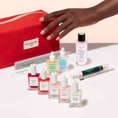 A hand reaches toward a red Manucurist Active™ Super Ultimate Manicure Kit bag and an array of manicure essentials—including nail polishes, remover, file, cuticle remover, and serum bottles—all beautifully arranged on a white surface for healthy nails.