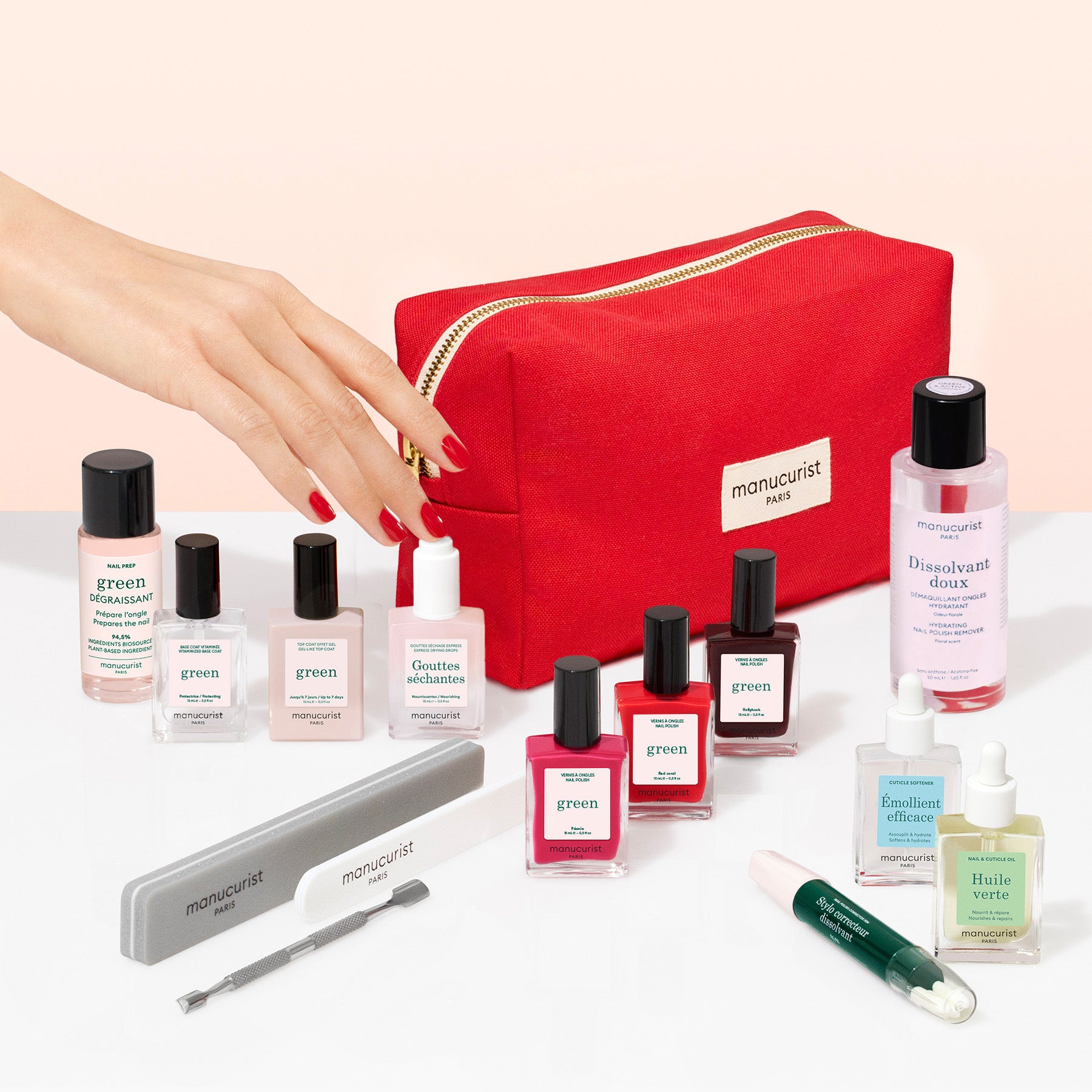 The Manucurist Addict Kit is displayed in a vibrant red bag, surrounded by a selection of nail care essentials including gel effect polishes, nail files, and treatment bottles, while a hand holds a polish brush close to the kit—everything you need for the perfect at-home manicure.