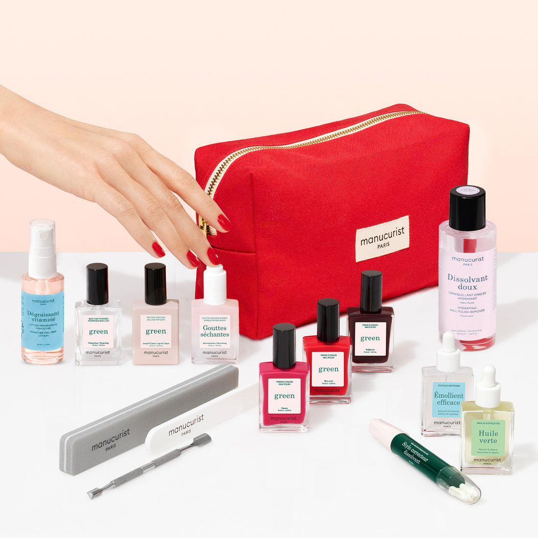 A hand reaches for a vitaminised base coat from the Addict Kit, surrounded by gel-effect nail polish bottles, remover, oil, a nail file, a metal cuticle pusher, a corrector pen, and a red zippered cosmetic bag.