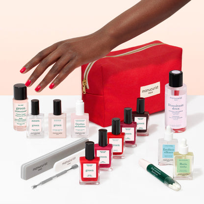 A hand with red nail polish reaches toward an open red cosmetics bag, surrounded by an array of nail care essentials—including polish bottles, files, oils, remover, and treatment bottles—neatly arranged on a white surface for the ideal at-home manicure with your favorite Manucurist Super Addict Kit.
