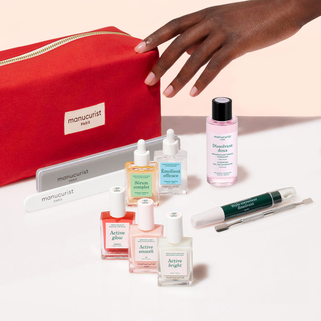 A hand reaches toward the Manucurist Active™ Ultimate Manicure Kit—a red nail care set featuring files, serums, polish bottles, cuticle remover, and a cuticle pen—each product designed to promote healthy nails and elegantly displayed on a light background.