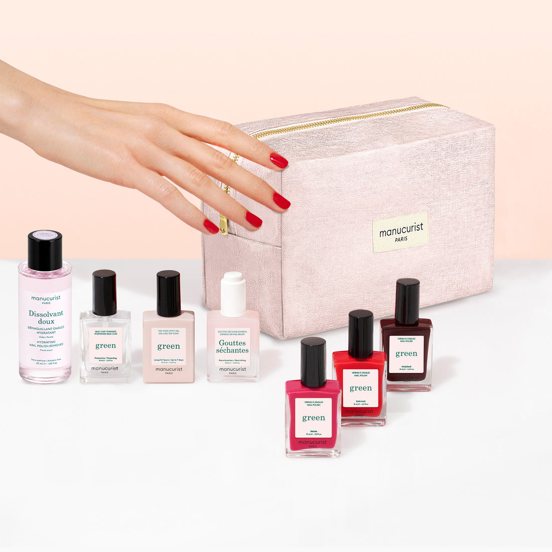 A hand with red nail polish reaches toward a pink Manucurist Starter Kit cosmetic bag, surrounded by various bottles of Manucurist gel-effect nail polish and nail care products arranged in front of the bag.