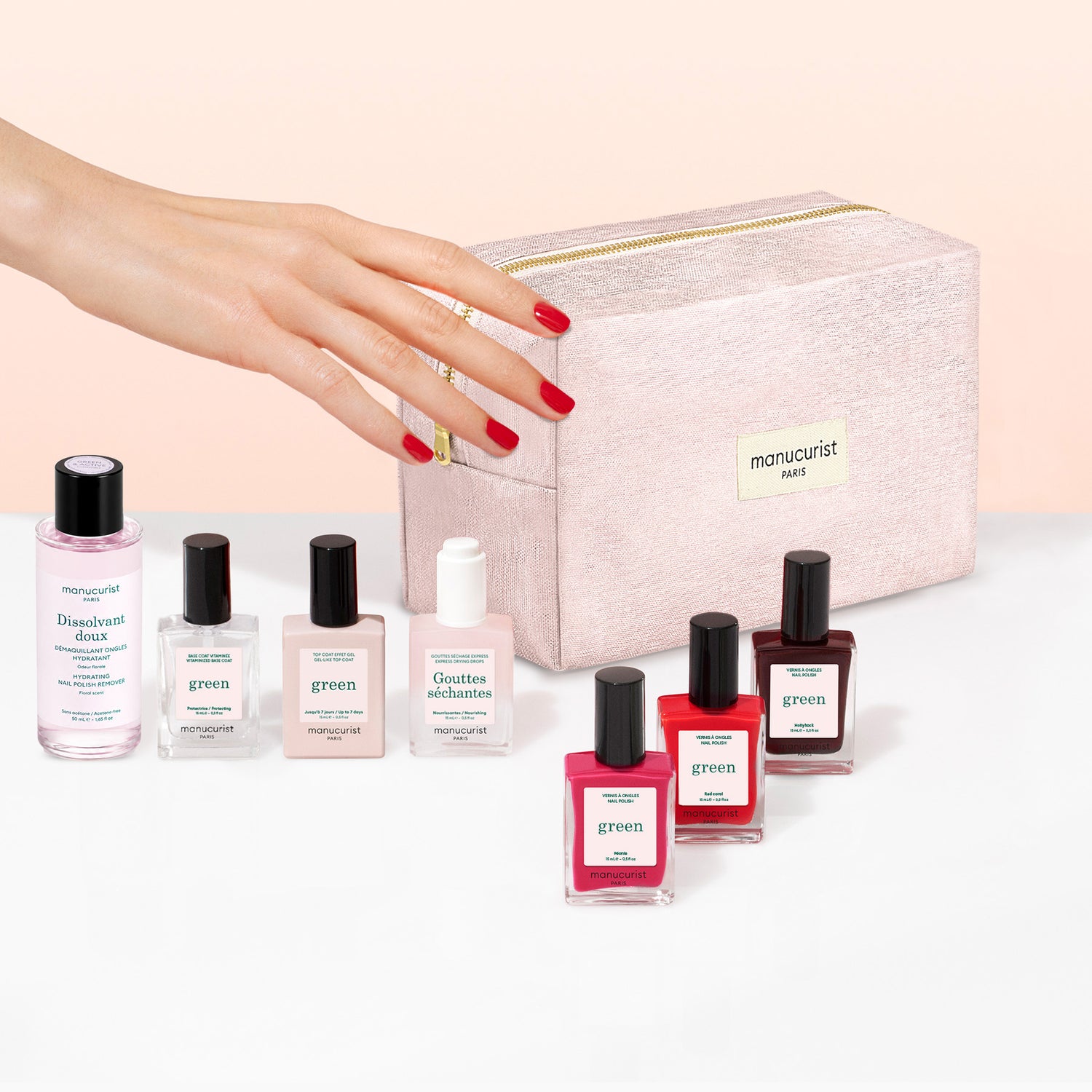 A hand with red nail polish reaches toward a pink Manucurist Starter Kit cosmetic bag, surrounded by various bottles of Manucurist gel-effect nail polish and nail care products arranged in front of the bag.