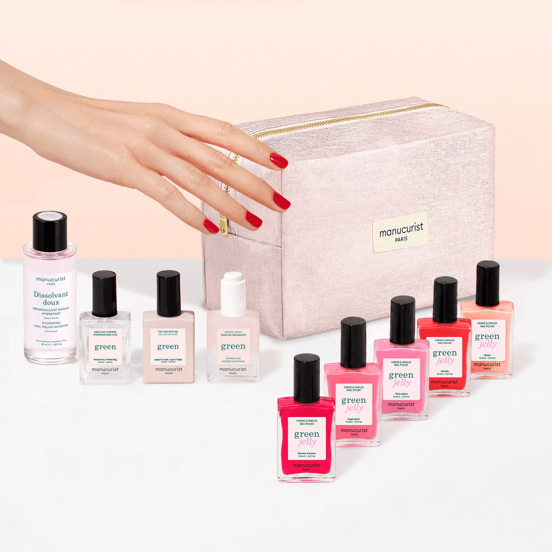 A hand with red nails reaches toward the Manucurist Super Starter Kit - Jelly Nail Polish, featuring a pink manicure set bag and an organic cotton pouch, surrounded by various pink and red jelly nail polishes, gel-effect polish, a remover bottle, and essential manicure tools.