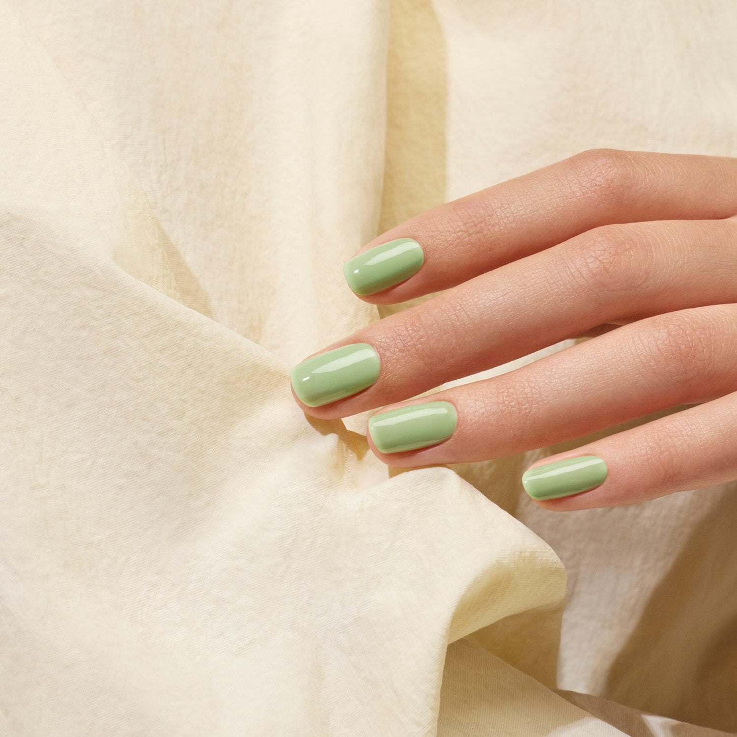 A hand with neatly manicured nails painted in Manucurist’s Milky Matcha nail polish from The Green range rests on a soft, cream-colored fabric background, exuding subtle botanical notes.