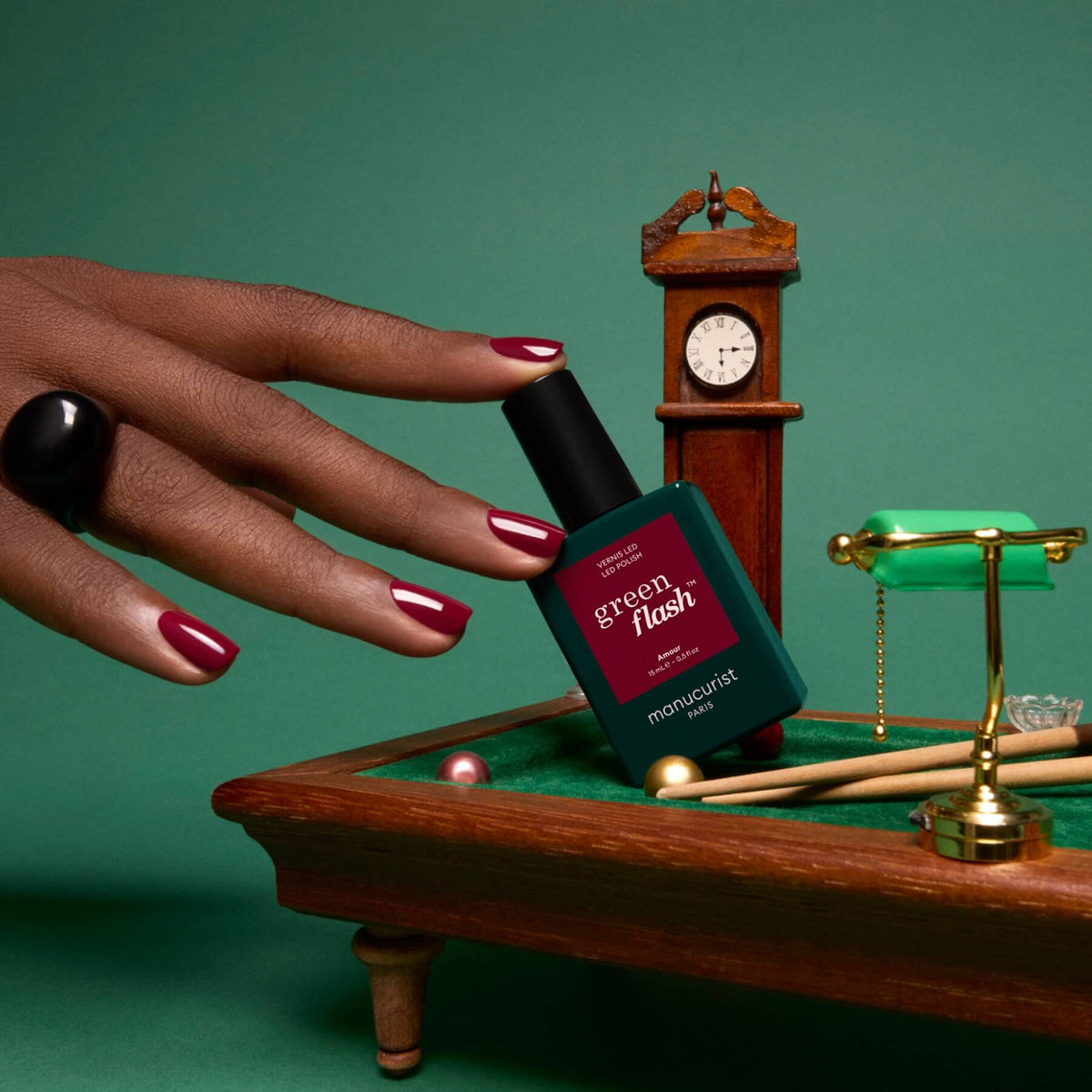 A hand with dark red nails, painted in the luminous burgundy shade “Amour” by Manucurist, holds a Green Flash gel polish bottle near a pool table, with a miniature grandfather clock and pool accessories in the background, set against a rich green backdrop.