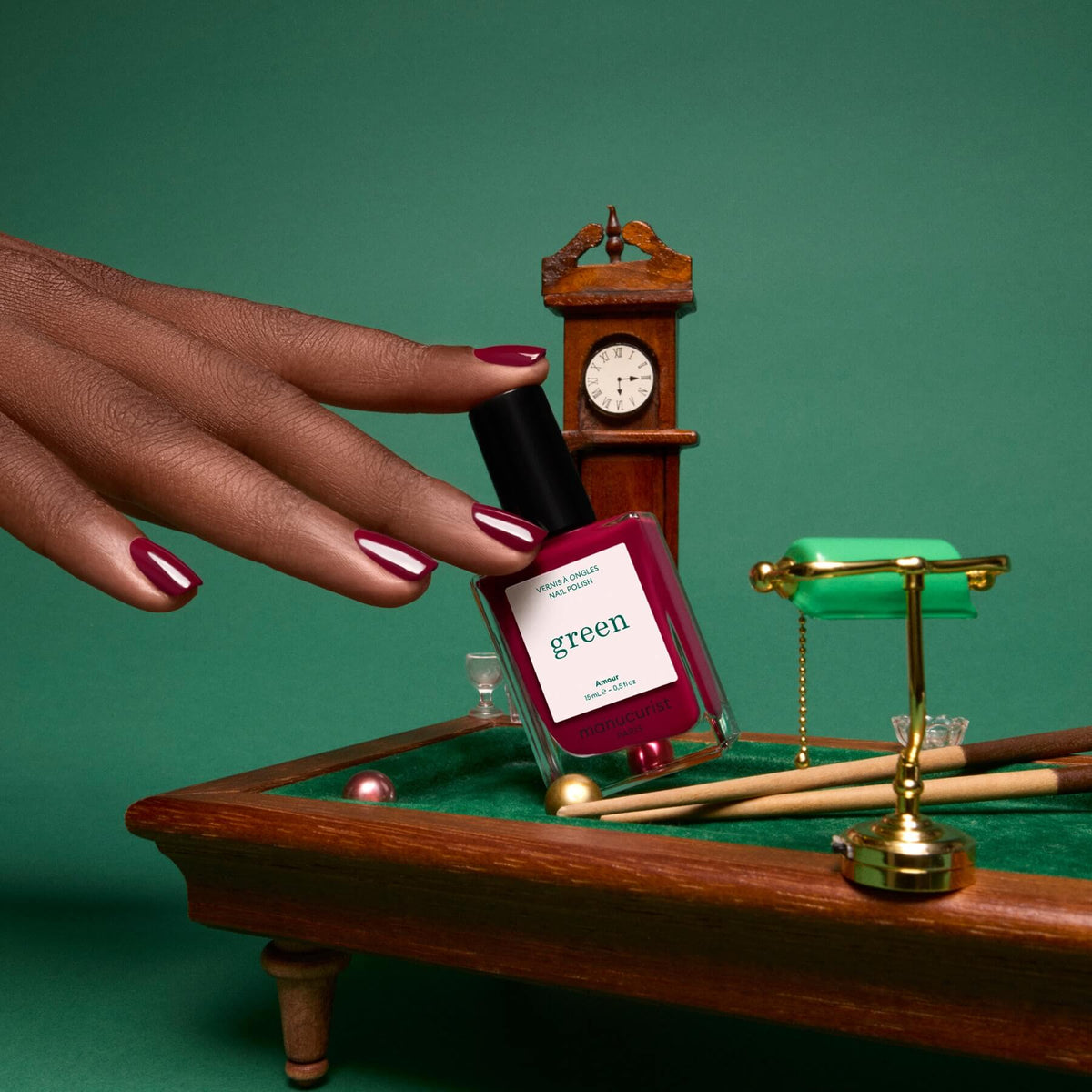 A hand with luminous burgundy Amour nail polish held over a miniature pool table and a small grandfather clock, hovering above a glass bottle of matching deep red nail polish.