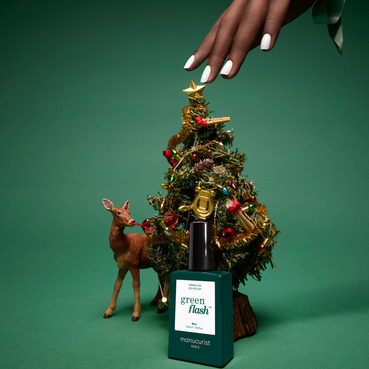 A hand with manicured nails decorates a small Christmas tree next to a deer figurine and a bottle of Manucurist Green Flash gel polish in the shade Bliss, all set against a green background. The shimmering finish of the iridescent white Bliss nail polish by Manucurist adds an elegant touch to the festive scene.