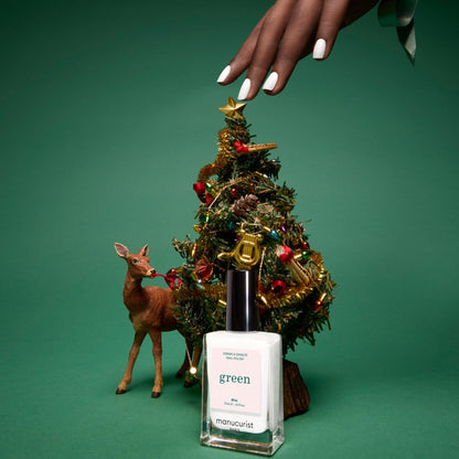 A hand with manicured nails reaches toward a small decorated Christmas tree beside a reindeer figurine. In front of them is a bottle of Bliss, the opaque pearly white nail polish from Manucurist.