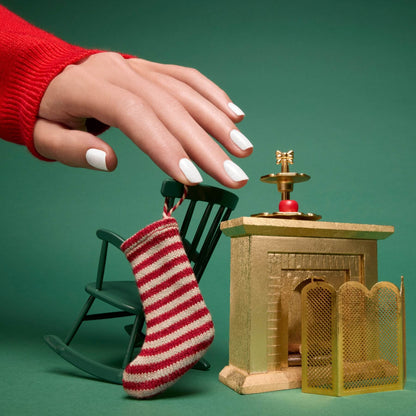A hand adorned with Bliss gel polish by Manucurist rests on a small green rocking chair beside a miniature gold fireplace featuring a striped stocking and mesh screen, set against a green background.