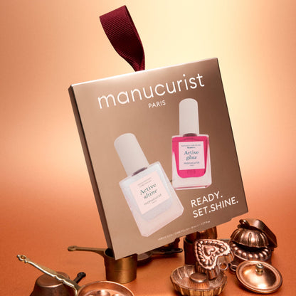 A box of Manucurist Active™ Travel-Size Duo nail polish set, featuring two bottles, is displayed among metallic decorative items against a warm copper-toned background. Ideal for nail care enthusiasts, this festive set adds a celebratory touch to your manicure routine.