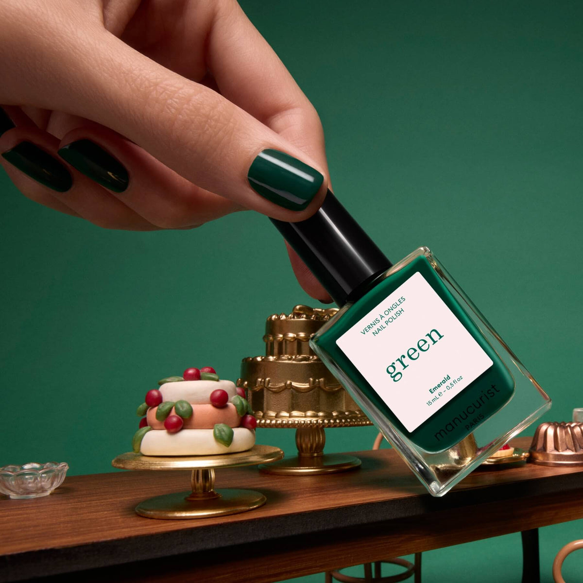 A hand with deep forest green-painted nails holds a bottle of Manucurist’s Emerald Green Flash LED gel nail polish above a table adorned with miniature cakes and desserts, set against a gemstone-inspired green background.