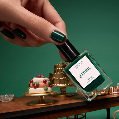 A hand with deep forest green-painted nails holds a bottle of Manucurist’s Emerald Green Flash LED gel nail polish above a table adorned with miniature cakes and desserts, set against a gemstone-inspired green background.