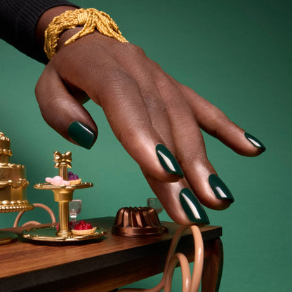 A hand with glossy, gemstone-inspired deep forest green nails—painted with Manucurist’s Green Flash LED gel nail polish in Emerald—hovers over a wooden table set with miniature cakes and pastries. The person wears a chunky gold bracelet, and the rich green background perfectly complements the striking shade of Emerald.