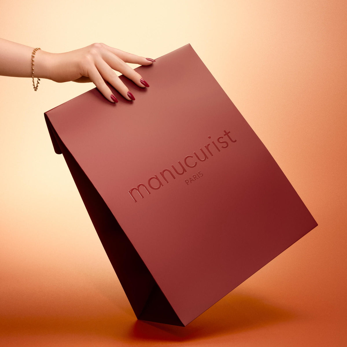 A hand with polished nails holds the Manucurist Holiday Gift Bag - M against a warm gradient background.
