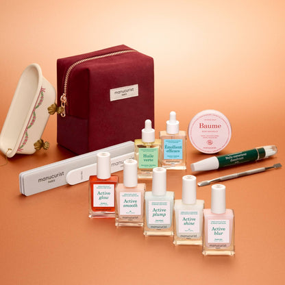 A burgundy zippered pouch containing a Manucurist Active™ Super Ultimate Kit Gift Set with five bottles of nail polish, cuticle oil, a dropper bottle, a small jar of balm, a nail file, and a cuticle pusher for a complete manicure routine.