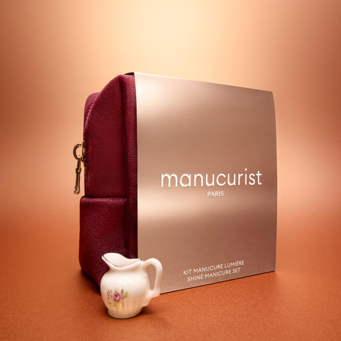 The Manucurist Active™ Gift Set Holiday, featuring a burgundy pouch and a beige Shine Manicure Set box, is artfully arranged alongside a small porcelain creamer with a floral design on a peach-colored surface against a warm gradient background.