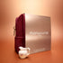The Manucurist Active™ Gift Set Holiday, featuring a burgundy pouch and a beige Shine Manicure Set box, is artfully arranged alongside a small porcelain creamer with a floral design on a peach-colored surface against a warm gradient background.