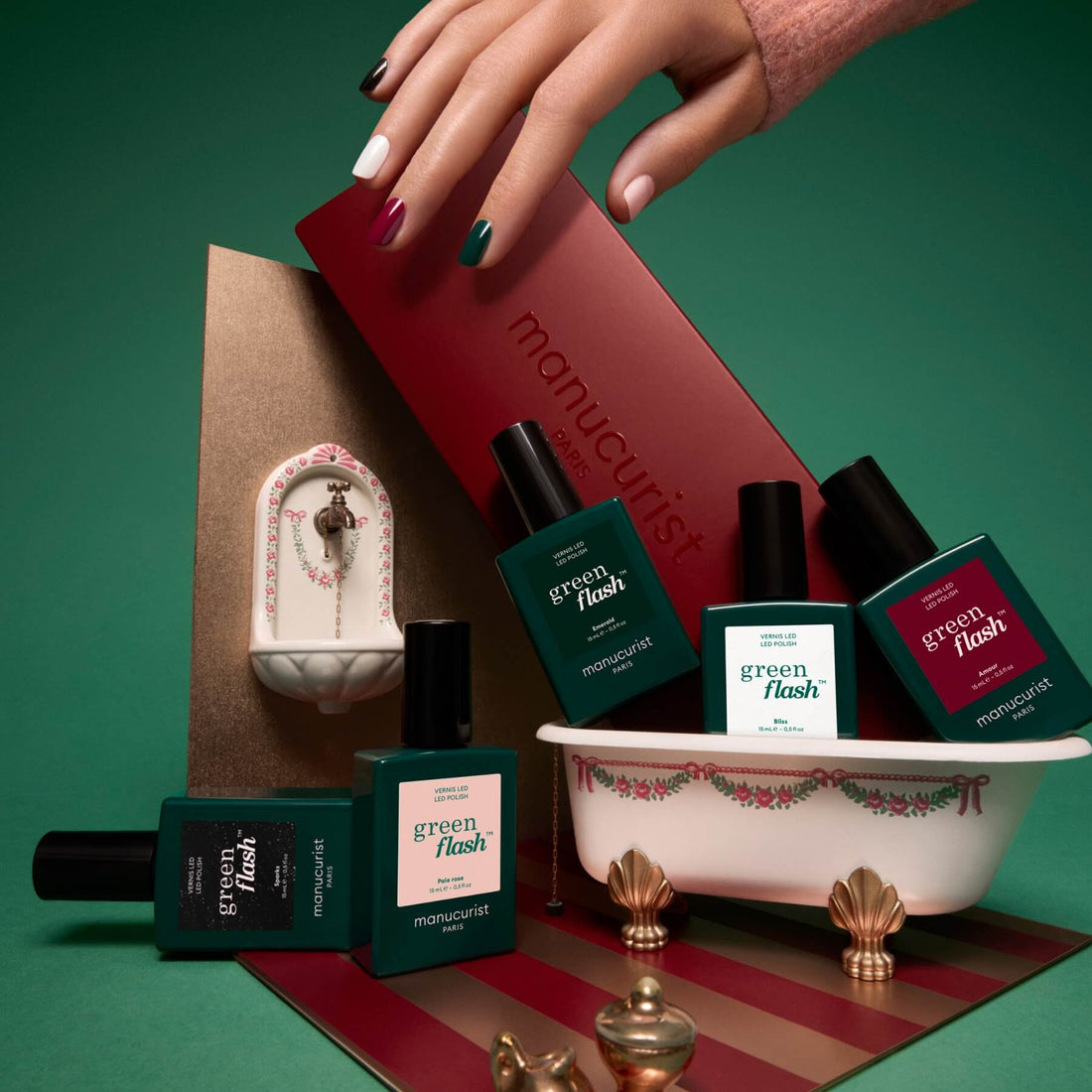A hand with multicolored nails holds a red box labeled Manucurist, perfectly showcasing the vibrant Rainbow Collections ideal for creative manicures. Nail polish bottles from Manucurist’s Rainbow Collections are displayed in a small decorative bathtub, with a tiny wall-mounted faucet in the background.