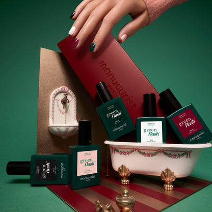 A hand with multicolored nails holds a red box labeled Manucurist, perfectly showcasing the vibrant Rainbow Collections ideal for creative manicures. Nail polish bottles from Manucurist’s Rainbow Collections are displayed in a small decorative bathtub, with a tiny wall-mounted faucet in the background.