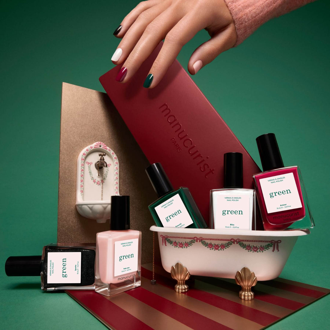 A hand with colorful manicured nails holds a red box above a small decorative bathtub filled with bottles of Manucurist’s Rainbow Collections, showcasing the latest nail trends and curated colour palettes from Manucurist.