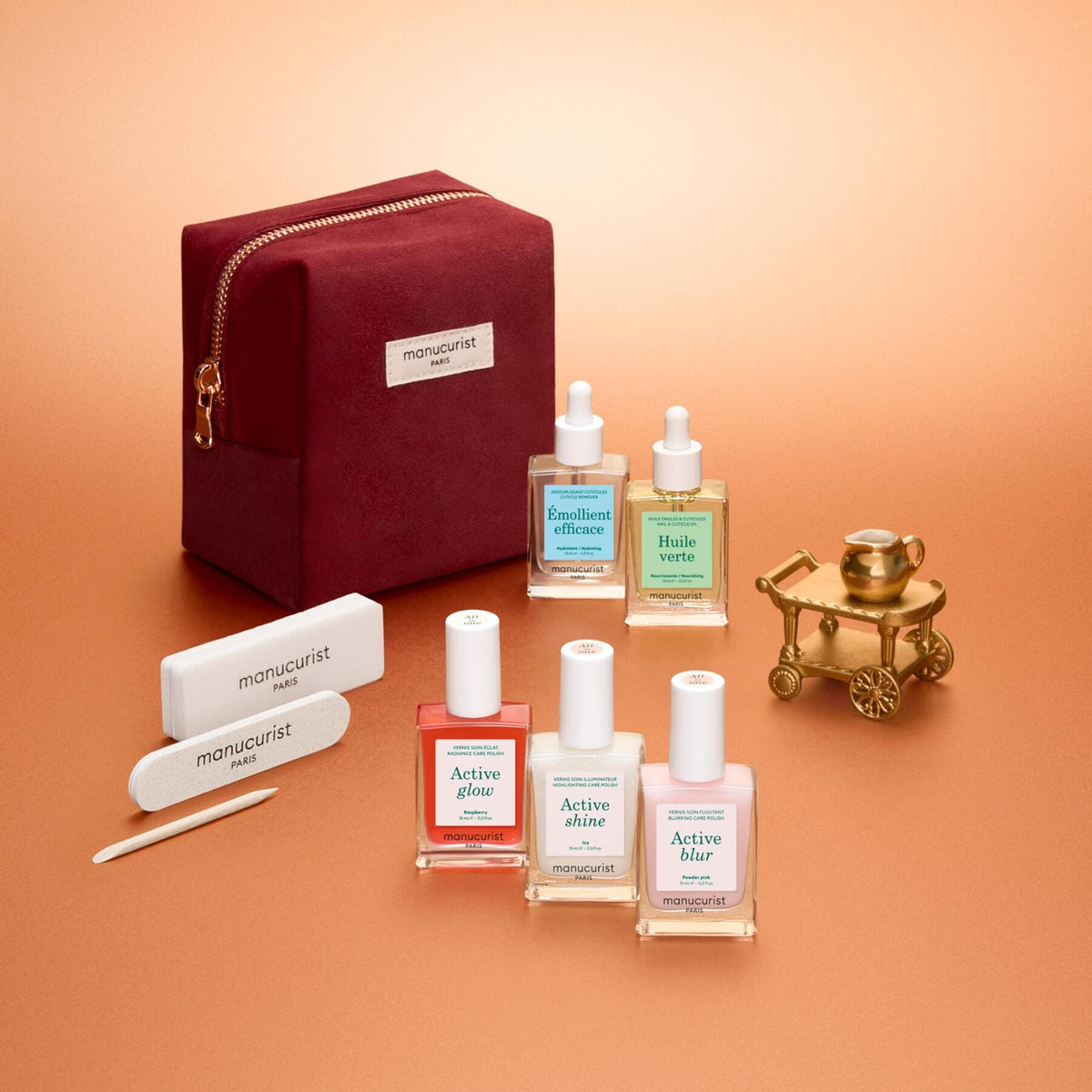 The Manucurist Active™ Starter Gift Set, featuring a burgundy pouch, nail care bottles with droppers, three nail files, and a gold miniature carriage, is beautifully arranged on a peach-colored surface—providing everything you need for a flawless manicure routine and beautiful nails.
