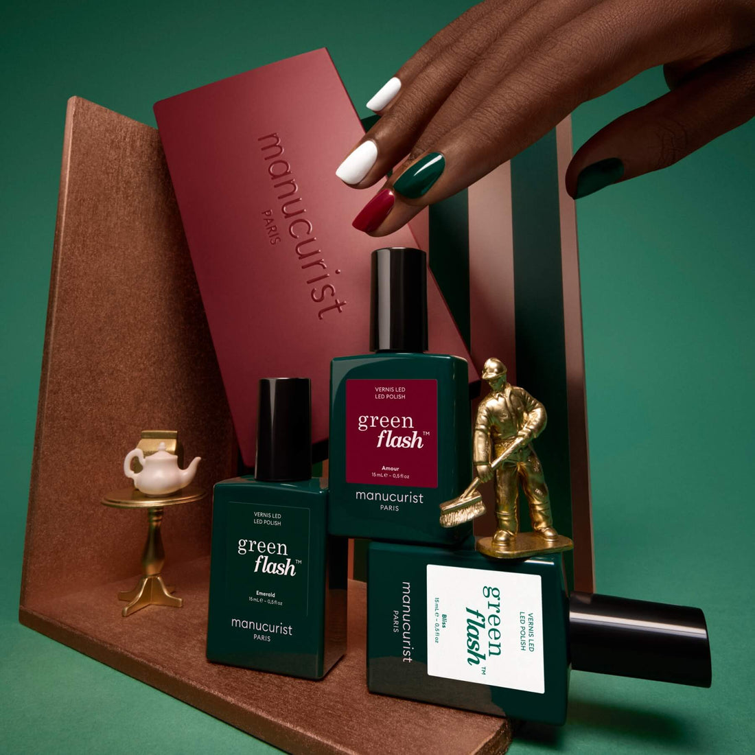 A hand with multicolored nails—showcasing the latest nail trends and vibrant colour palettes—reaches toward green nail polish bottles from the Trio Collections by Manucurist, arranged alongside a teapot, books, and a small hockey player statue on a green background.