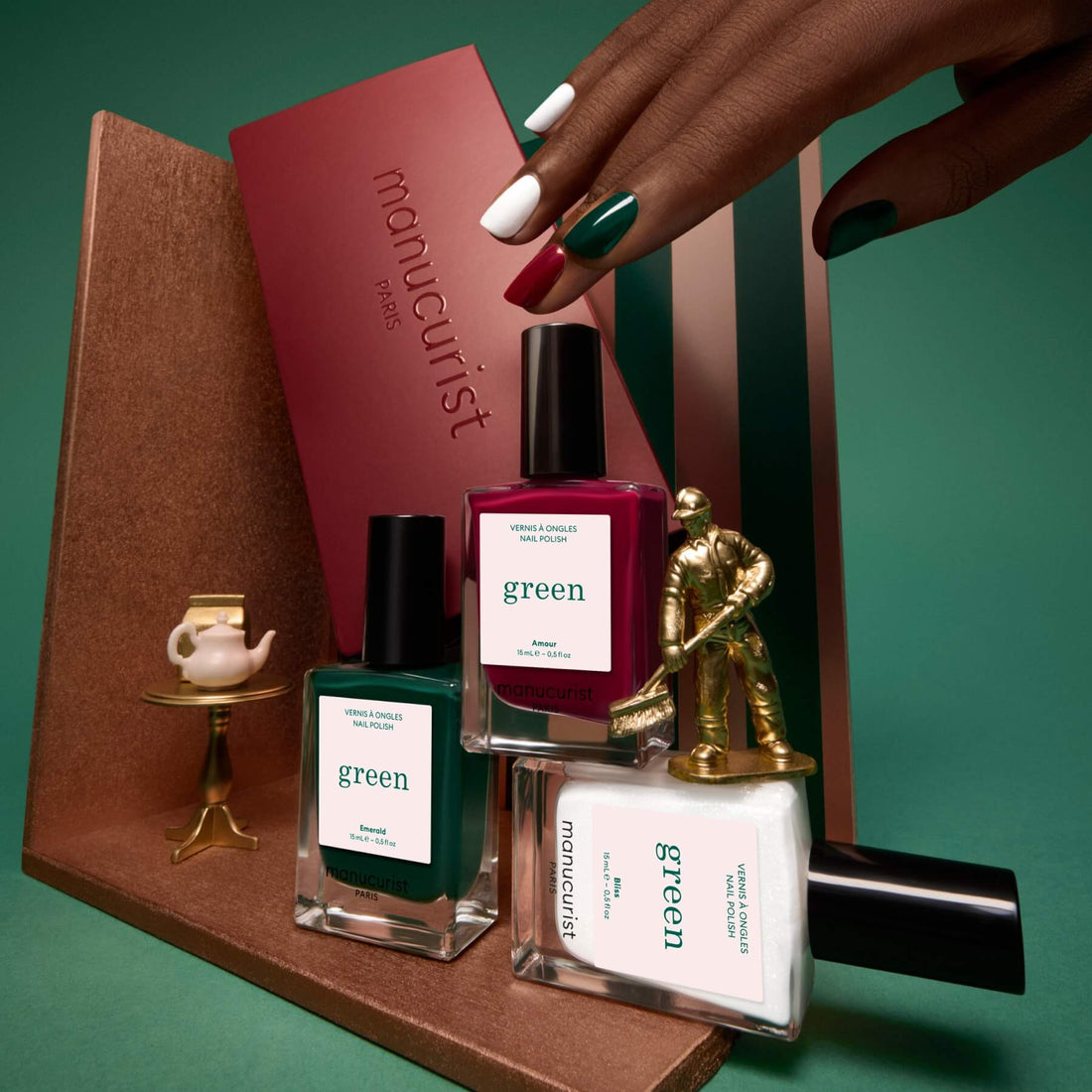 A manicured hand holds a burgundy box from Manucurist’s Trio Collections beside green, white, and burgundy nail polish bottles, all arranged on a brown display with a tiny gold statue and a cup against a green background—perfect inspiration for anyone seeking the latest nail trends and curated colour palettes.