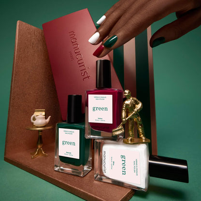 A manicured hand holds a burgundy box from Manucurist’s Trio Collections beside green, white, and burgundy nail polish bottles, all arranged on a brown display with a tiny gold statue and a cup against a green background—perfect inspiration for anyone seeking the latest nail trends and curated colour palettes.