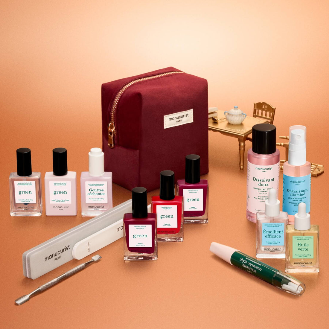 A Green™ Addict Kit Gift Set featuring a burgundy makeup bag surrounded by naturally derived nail polishes, skincare bottles, a nail file, and a cuticle pusher on a peach surface for an at-home manicure.