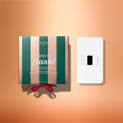 A green and pink striped Manucurist Advent Calendar Time to Flash! box sits next to a small white LED lamp with buttons and a screen, both displayed on a peach-colored background—perfect for creating your next gel polish manicure.