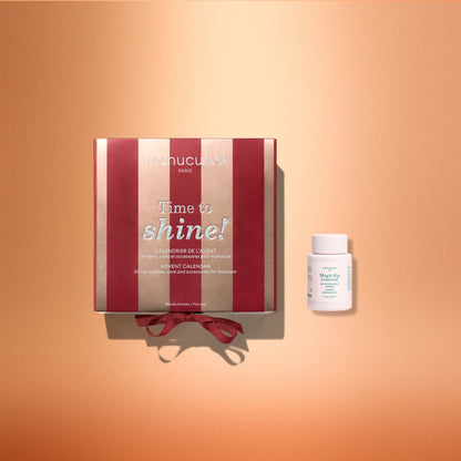 A red and gold striped box labeled “Advent Calendar Time to Shine!” from Manucurist rests on a peach background beside a small white bottle of Magic Remover nail polish remover, creating an inviting scene for nail care enthusiasts.