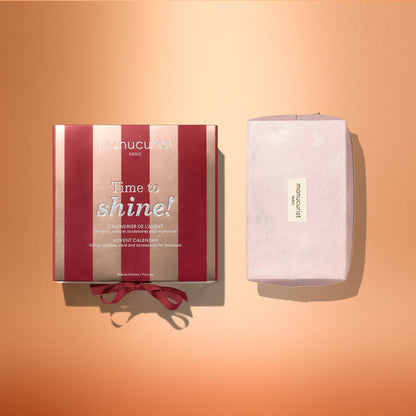 A red and pink striped Manucurist Advent Calendar Time to Shine! box, featuring the phrase “Time to shine!”, sits beside a pink-wrapped gift on a peach gradient background, inviting you to unveil icy Active Shine nail care surprises inside.