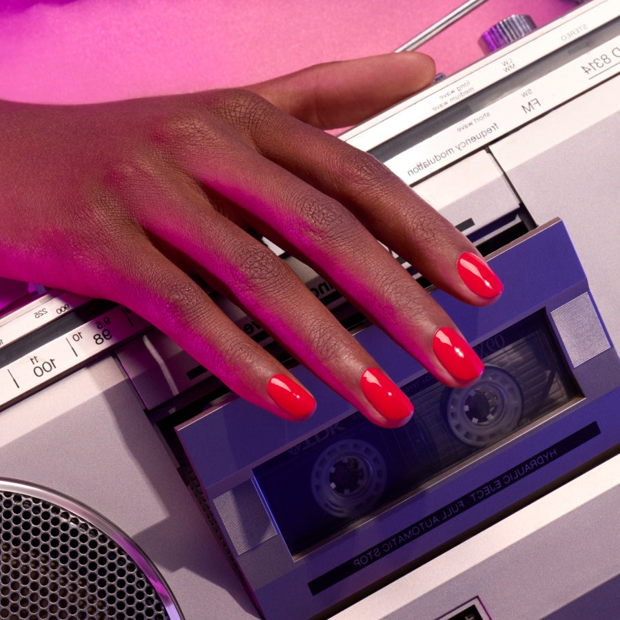 A hand with Manucurist’s Spicy nail polish, a vibrant red gel shade, rests on a retro cassette tape player under pink lighting.